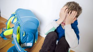 Ein Junge sitzt traurig in seinem Kinderzimmer neben seinem Schulranzen und versteckt sein Gesicht hinter seinen Haenden.