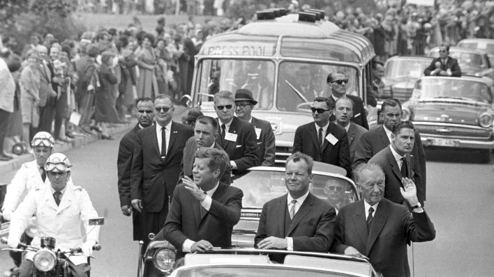 Archivbild: John F. Kennedy mit dem Regierenden Bürgermeister Willy Brandt (M) und Bundeskanzler Konrad Adenauer (r) während der Fahrt im offenen Wagen durch Berlin. (Quelle: dpa)