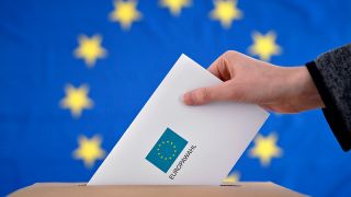 Symbolbild: Ein Wahlbrief wird in eine Wahlurne gesteckt, im Hintergrund eine EU-Flagge. (Quelle: dpa/Panama)