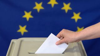 Symbolbild: Eine Hand steckt einen Umschlag in eine Wahlurne vor der Europafahne. (Quelle: dpa/Ending)