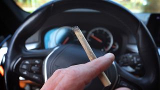 Symbolbild: Ein Mann sitzt mit einem Joint zwischen den Fingern am Steuer eines Autos. (Quelle: dpa/Karl-Josef Hildenbrand)