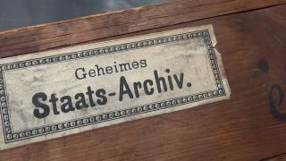Es ist eine Holzkiste zu sehen auf der eine Plakette angebracht ist auf welcher steht: "Geheimes Staats-Archiv".