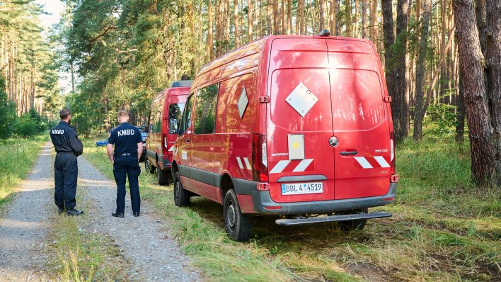 Zwei Mitarbeiter des Kampfmittelbeseitigungsdienstes stehen am 29.06.2024 neben einem Fahrzeug im Wald nahe der Tesla-Fabrik. Geplant ist die Sprengung einer 250-Kilo-Bombe. (Quelle: dpa-Bildfunk/Annette Riedl)