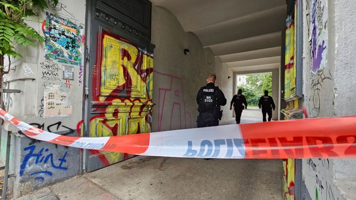 In einem mit Flatterband abgesperrten Hauseingang gehen drei Polizisten. Nach Störaktionen an mehreren Flughäfen in Deutschland durchsucht die Polizei Wohnungen von Mitgliedern der Klimagruppe Letzte Generation. (Quelle: dpa/Junghannß)