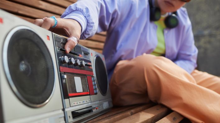 Symbolbild:Eine Person sitzt auf einer Bank und drückt einen Knopf auf der Boombox.(Quelle:imago images/Pond5 Images)