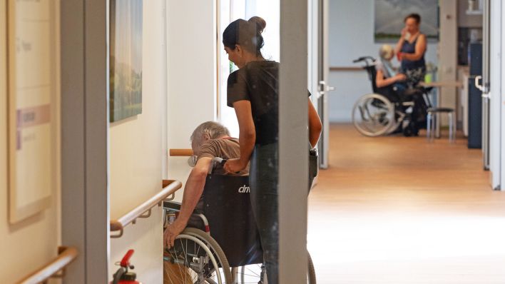 Symbolbild: Eine Pflegerin schiebt einen Patienten im Rollstuhl am 29.08.2024. (Quelle: picture alliance/SvenSimon/Malte Ossowski)