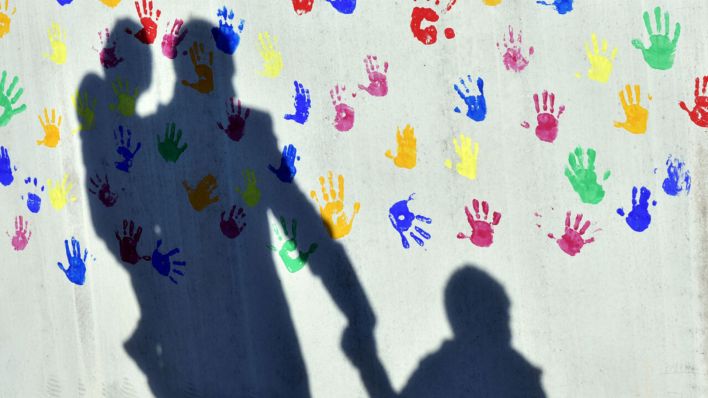 Symbolbild: Ein Mann mit einem Kind auf dem Arm und einem an der Hand wirft einen Schatten auf eine mit bunten Handabdrücken bemalte Wand einer Kindertagesstätte. (Quelle: dpa/Kneffel)